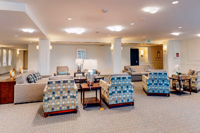 A spacious and well-lit living room area with multiple patterned armchairs and sofas arranged around wooden side tables with lamps. The room has beige walls, carpeted floors, and ceiling lights, creating a comfortable and inviting atmosphere.