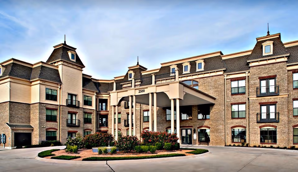 Front exterior view of a large, elegant senior living facility building with multiple windows, a covered entrance supported by columns, and well-maintained landscaping including bushes and flowers in front.