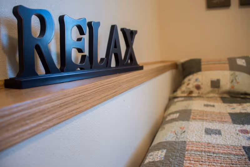 A close-up view of a wooden shelf with a decorative sign spelling 'RELAX' in large black letters. Next to the shelf is a bed with a patchwork quilt featuring various patterns and colors.
