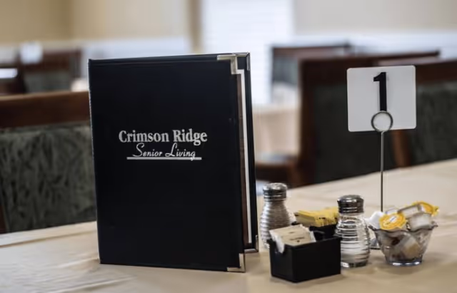 A dining table setup with a Crimson Ridge Senior Living menu, condiments, and a table number sign.