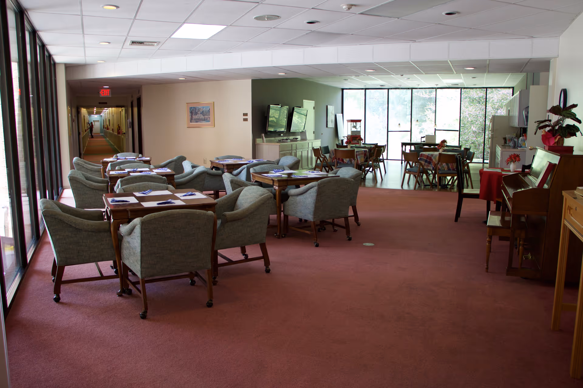 A spacious common area in a senior living facility with multiple seating arrangements including tables and cushioned chairs. The room has large windows letting in natural light, a piano on the right side, and a hallway leading to other rooms on the left. The carpet is a muted red color and the walls are light-colored.