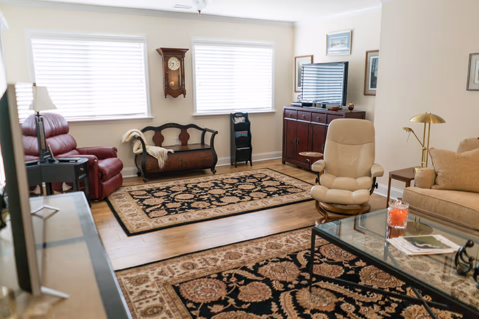 Well-lit living room with multiple seating options including a red recliner and beige swivel chair, patterned area rugs, a TV cabinet, and a glass coffee table.