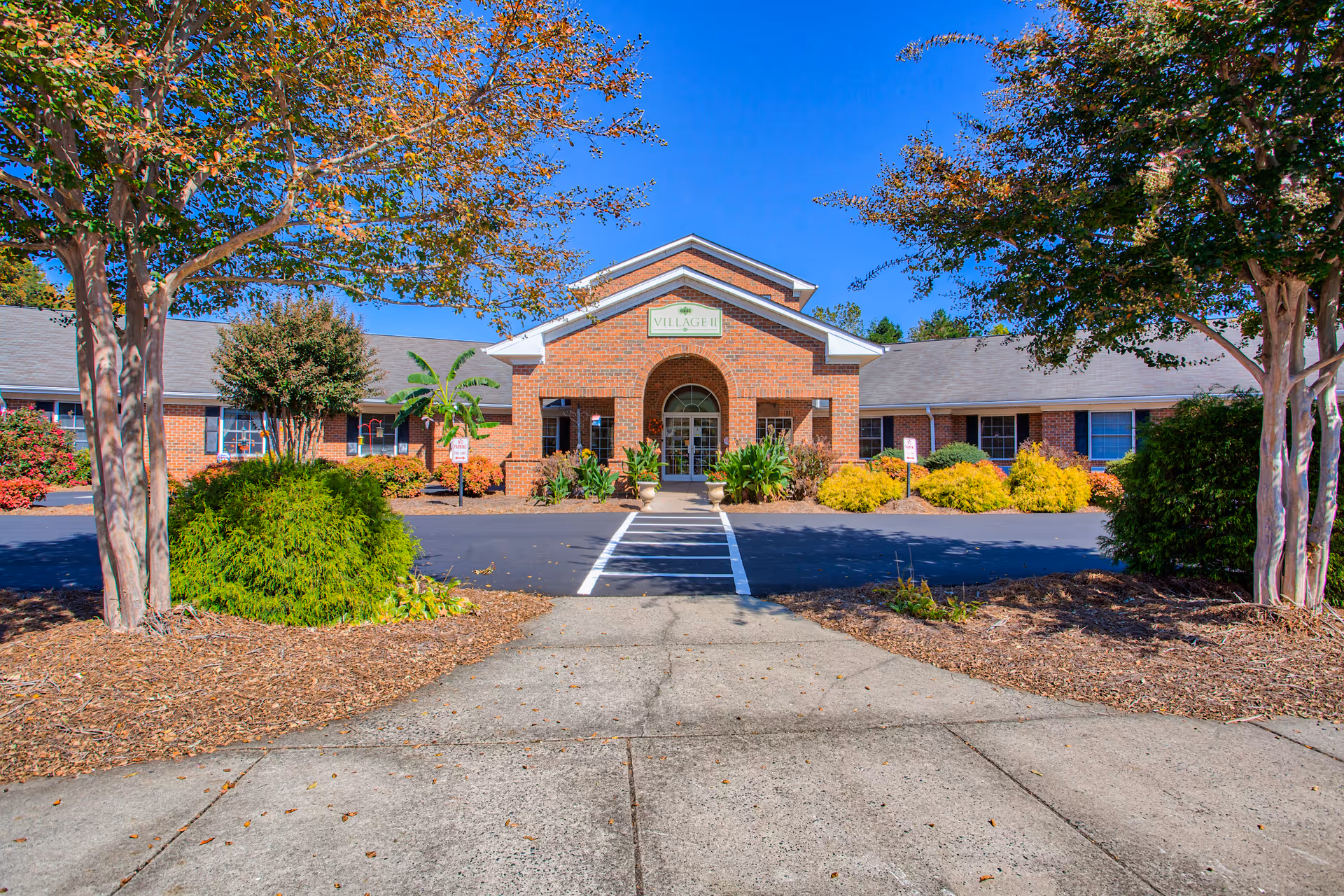 Front exterior view of Clemmons Village II- Assisted Living facility showing a brick building with a central entrance under a peaked roof. The entrance is framed by an archway and flanked by plants and shrubs. Trees and landscaped bushes line the driveway and walkway leading to the entrance under a clear blue sky.