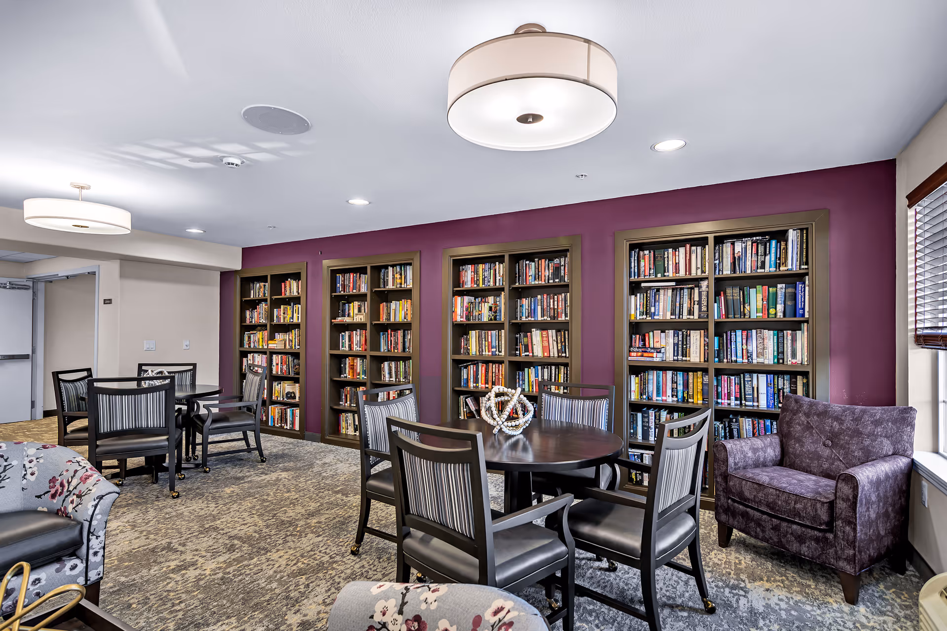 Communal library lounge with round tables and chairs, built-in bookshelves along a purple wall and a purple armchair by the window.
