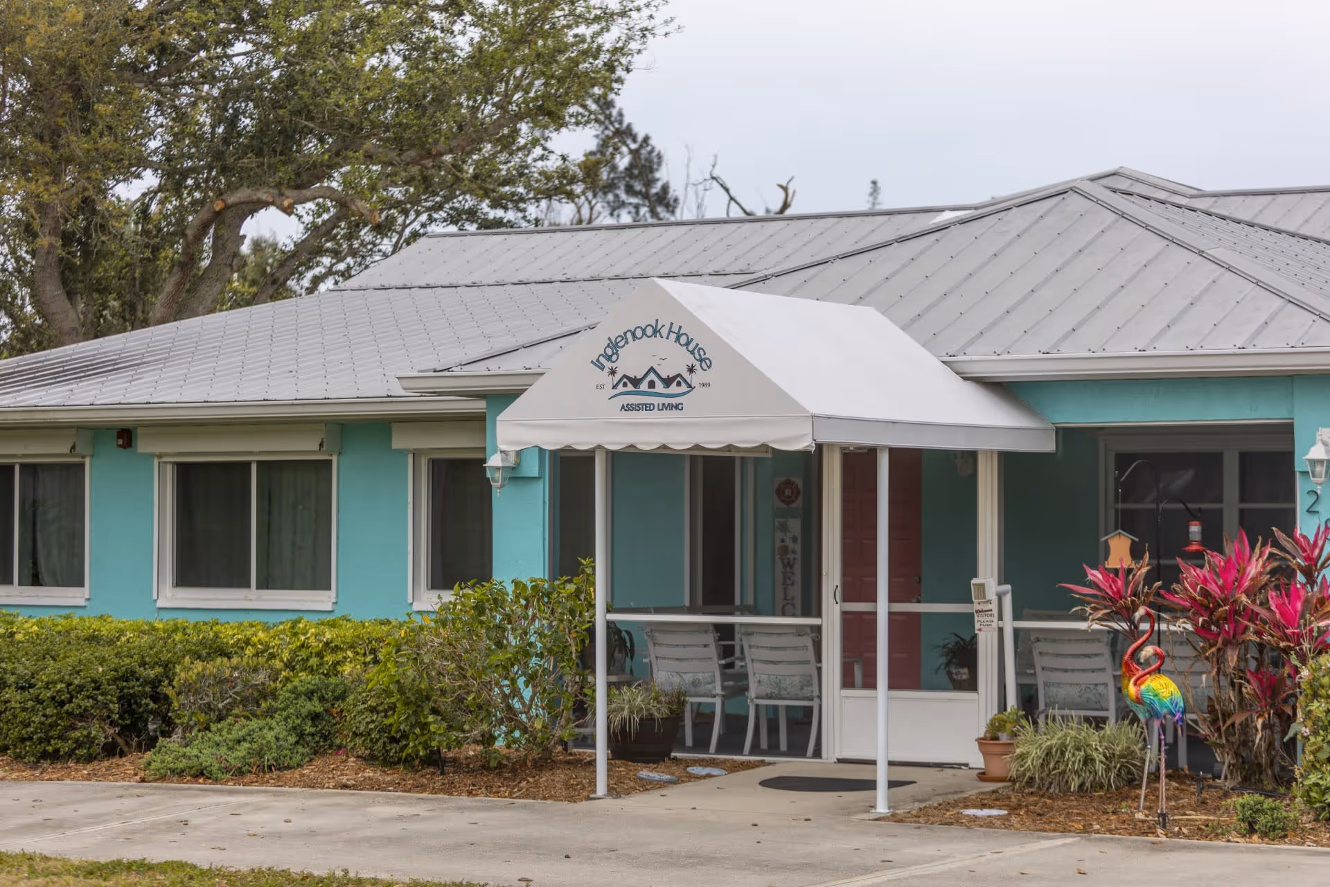Exterior view of Inglenook House Assisted Living & Memory Care building with a light blue facade, white metal roof, and a white awning over the entrance displaying the facility's name and logo. There are bushes and plants around the entrance, along with outdoor chairs and a colorful flamingo decoration.
