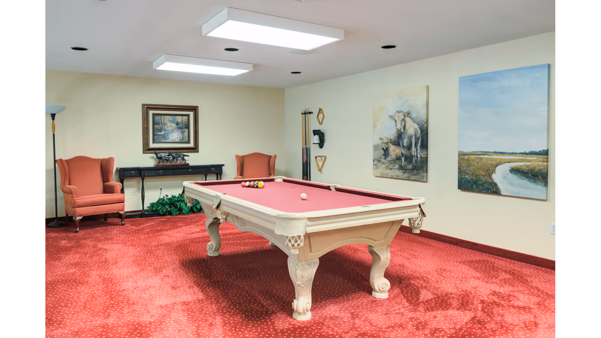 A recreation room interior featuring a white pool table with red felt on red carpeting, two upholstered chairs, wall art, and pool cues.