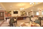 A spacious dining room with multiple round tables covered with white tablecloths and set with napkins and small centerpieces. The room features cushioned chairs with floral upholstery, a large wall-mounted television, and decorative wall art with floral patterns. The ceiling has recessed lighting and a chandelier, and there is a long counter or buffet area along one wall.