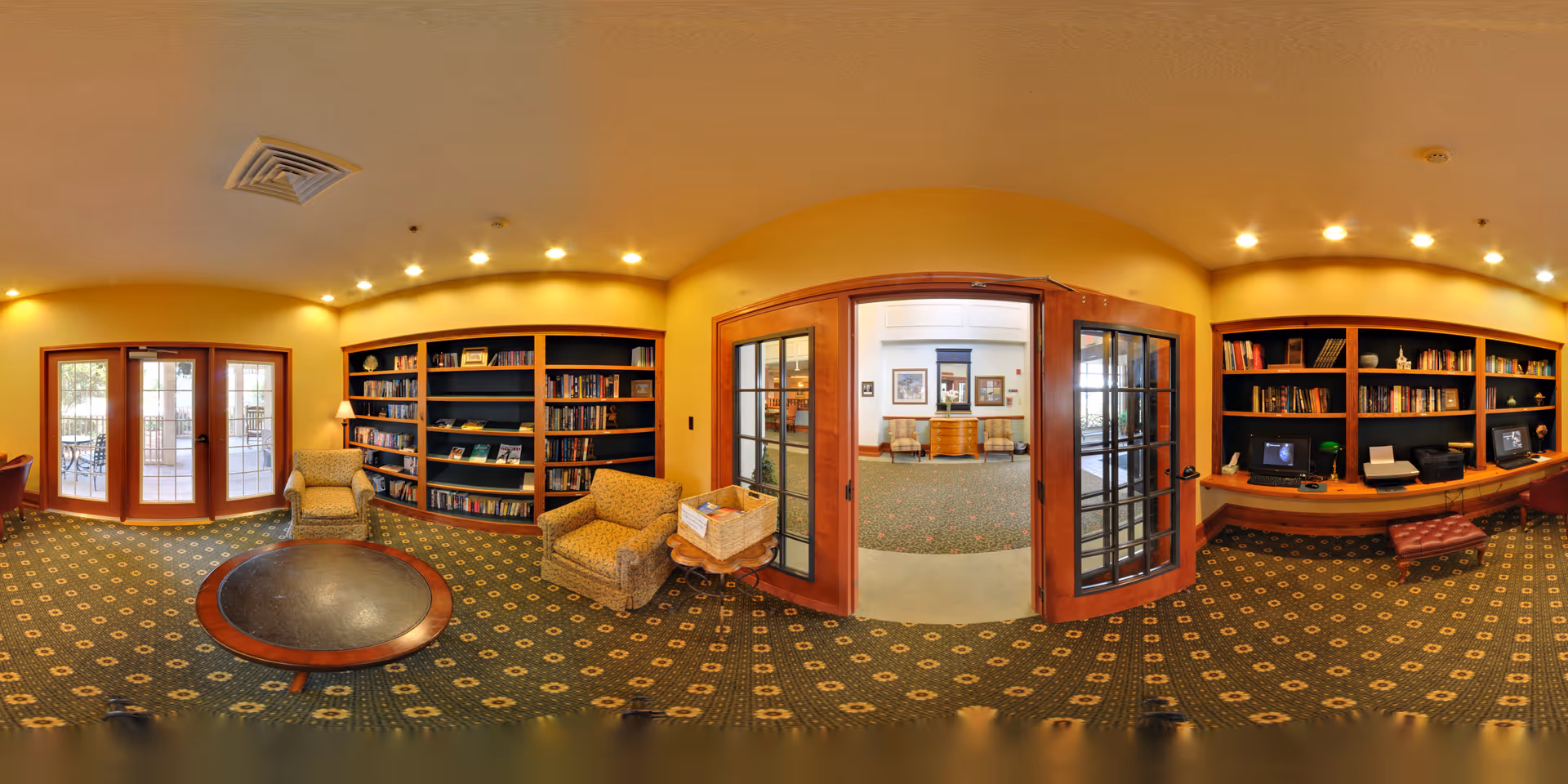 A cozy common area with yellow walls and patterned carpet. The room features wooden bookshelves filled with books and decorative items, two upholstered armchairs, a round wooden table, and a basket on a small table. There are glass double doors leading to another room with additional seating and framed pictures on the walls. On the right side, there is a desk area with two computer stations and a printer.