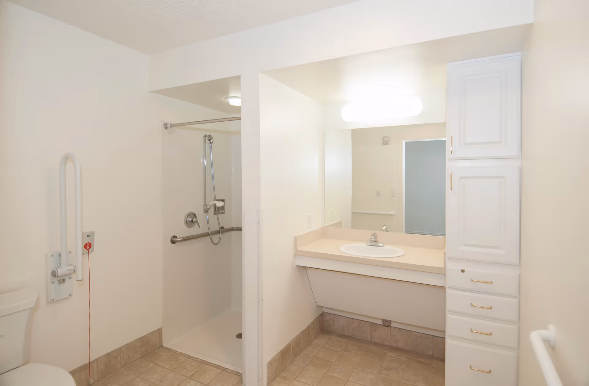 A clean and accessible bathroom with a walk-in shower featuring grab bars and a handheld showerhead. There is a white toilet with a grab bar and emergency pull cord next to it. The bathroom also has a large mirror above a sink with a beige countertop and white cabinetry with gold handles. The walls are painted white and the floor is tiled.