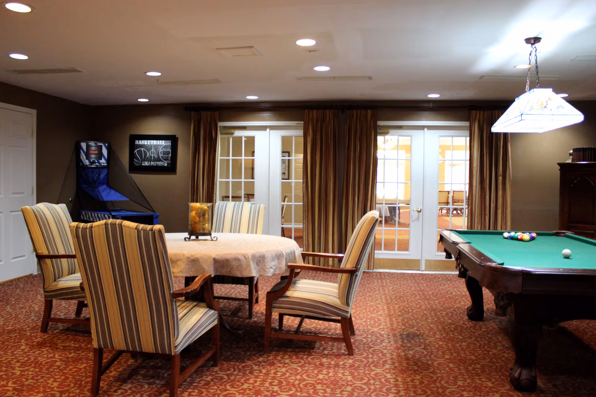 A recreational room with a round table covered with a white tablecloth surrounded by four striped upholstered chairs. To the right is a pool table with billiard balls arranged for a game. In the background, there are double glass doors with brown curtains and a basketball arcade game against the wall. The room has a patterned red carpet and recessed ceiling lights.