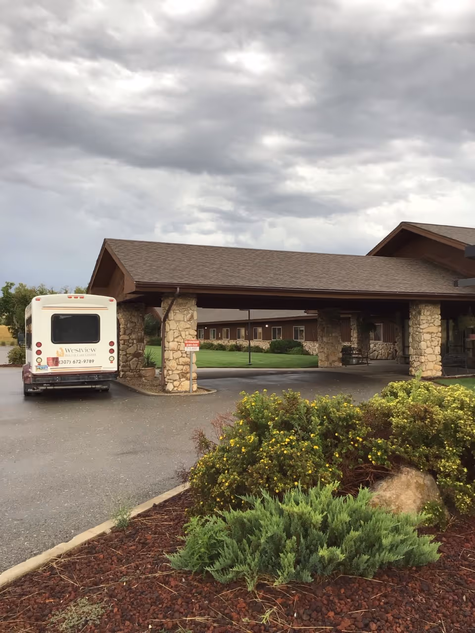 Covered entrance of Westview Health Care Center with a parked shuttle bus, stone pillars, and landscaped shrubs under a cloudy sky.