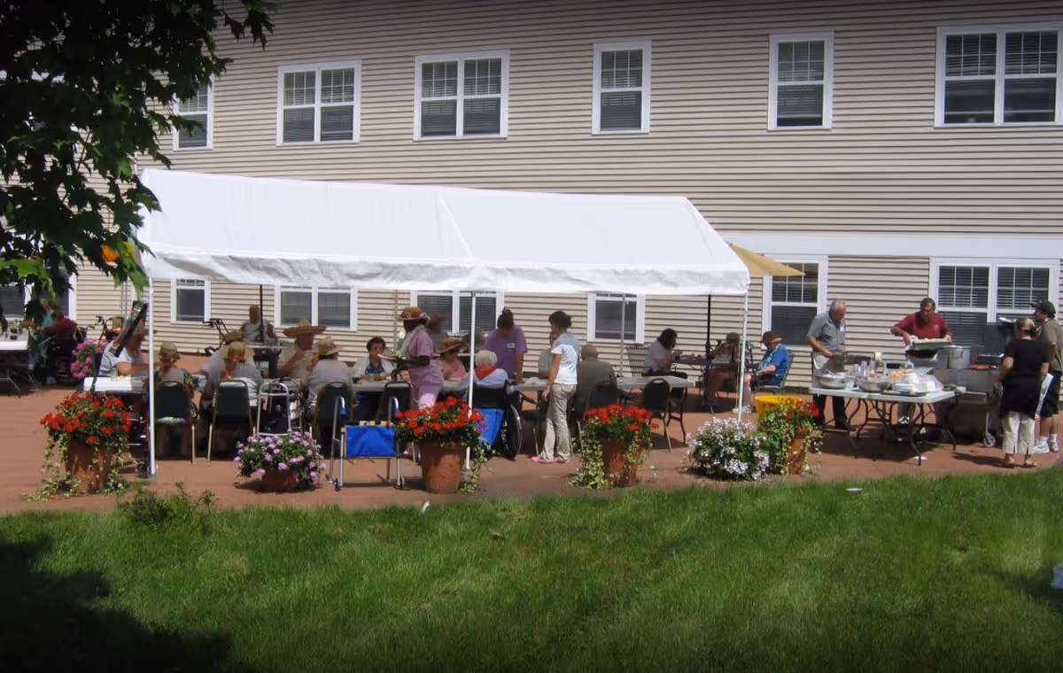 Outdoor patio area at an assisted living facility with a white canopy tent covering several elderly residents seated at tables. Staff members are assisting and serving food from a buffet table nearby. The building with multiple windows is visible in the background, and there are potted flowers decorating the patio.