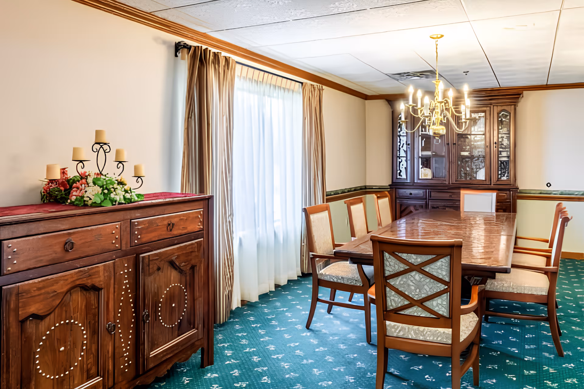 A formal dining room with a long wooden table surrounded by eight upholstered chairs. There is a wooden sideboard with a floral arrangement and candle holder on top, a large window with beige curtains, and a wooden china cabinet in the corner. A chandelier with lit bulbs hangs above the table, and the floor is covered with a green patterned carpet.