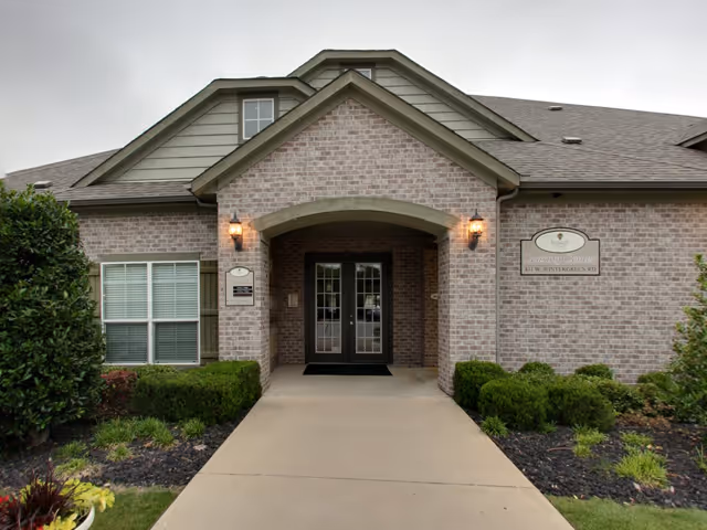 Front exterior view of a brick building with a covered entrance featuring double glass doors, two wall-mounted lantern lights, and surrounding greenery including bushes and small plants.
