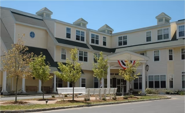 Exterior view of a multi-story senior living facility building with beige siding, multiple windows, and a covered entrance decorated with red, white, and blue bunting. There are small trees and white benches in front of the building along a paved driveway.