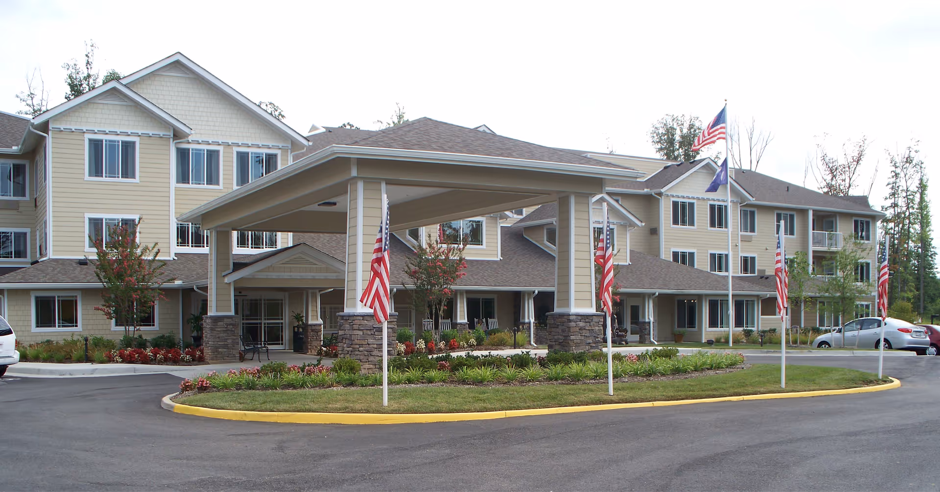 Exterior view of Chesterfield Heights Gracious Retirement Living facility showing a large, multi-story building with beige siding and multiple windows. The entrance has a covered drop-off area supported by columns with stone bases. Several American flags are displayed on flagpoles around the circular driveway, with landscaped greenery and flowers in the center.