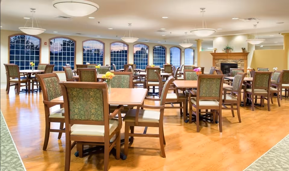 A spacious dining room with multiple wooden tables and chairs arranged neatly. Each table has a small vase with colorful flowers. Large arched windows line the far wall, allowing natural light to fill the room. The room features a wooden floor, soft lighting from ceiling fixtures, and a fireplace with a wooden mantel on the right side.
