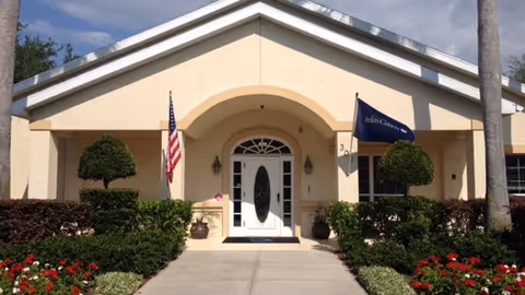 Front exterior view of a single-story building with a peaked roof, featuring a white door with decorative glass panels. The entrance is flanked by two potted plants and two trimmed bushes. An American flag is displayed on the left side, and a blue flag with the text 'Arden Courts' is on the right. The walkway leading to the door is bordered by green shrubs and red flowers.