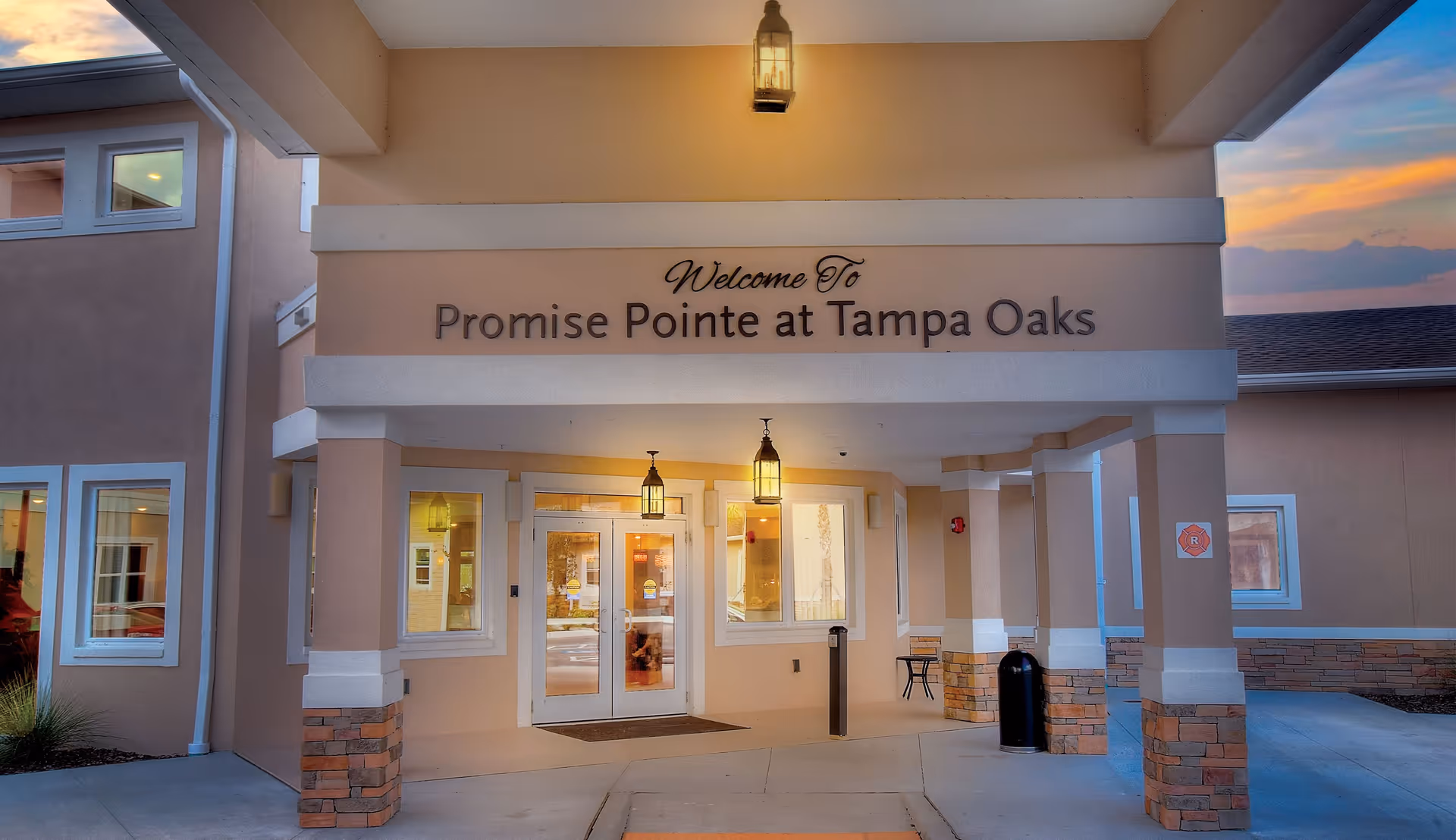 Entrance of Promise Pointe at Tampa Oaks senior living facility during sunset, featuring a covered entryway with hanging lantern lights, double glass doors, and beige exterior walls with white trim and brick accents.