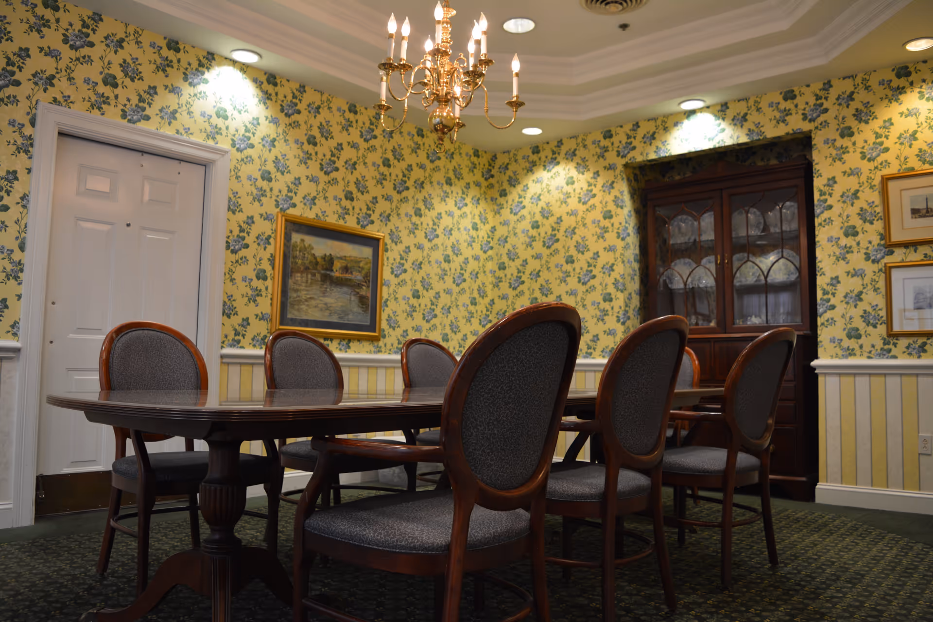 A traditional dining room with a dark wooden table surrounded by six upholstered chairs. The walls are covered with yellow floral wallpaper and decorated with framed paintings. A wooden china cabinet with glass doors is visible in the corner. A brass chandelier hangs from the ceiling, illuminating the room.