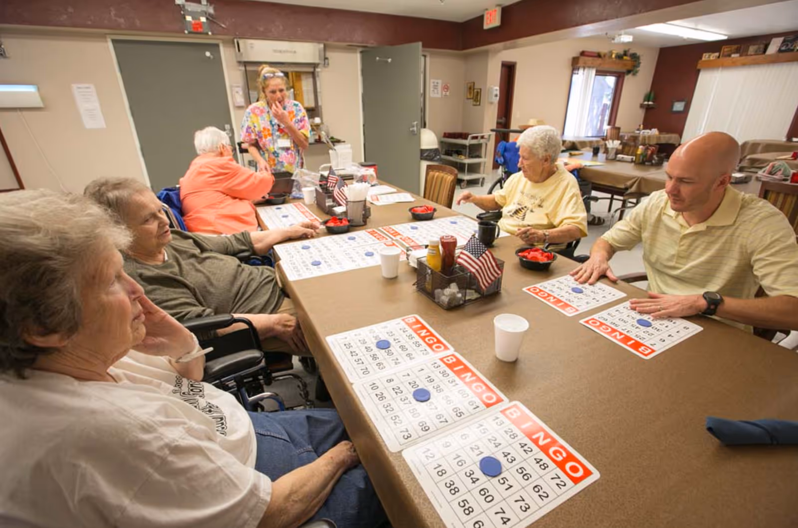 A group of elderly people and a man sitting around a table playing bingo in a communal room. A staff member stands nearby, observing and smiling. The table is covered with bingo cards, markers, cups, and small American flags. The room has a warm, welcoming atmosphere with chairs and tables in the background.
