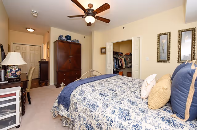 A bedroom with a bed covered in a blue and white floral patterned comforter and multiple pillows. The room has beige walls, a ceiling fan with a light, a wooden armoire, and an open closet with clothes hanging inside. There is a desk with a lamp and chair on the left side, and two framed floral pictures on the wall to the right.