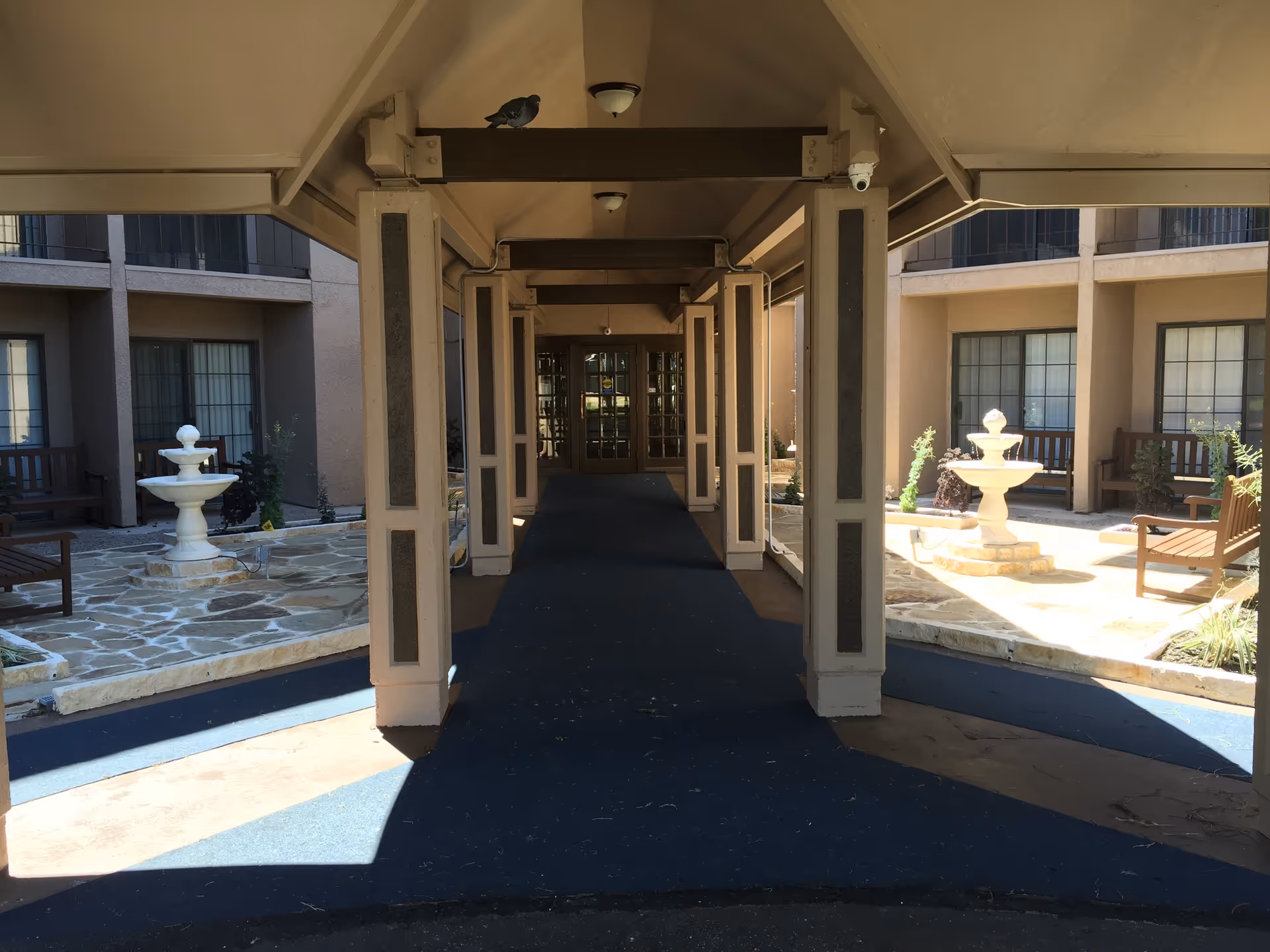 Covered walkway leading to a building entrance with glass doors, flanked by outdoor seating areas with benches and two white tiered fountains on stone patios. A pigeon is perched on a beam above the walkway.