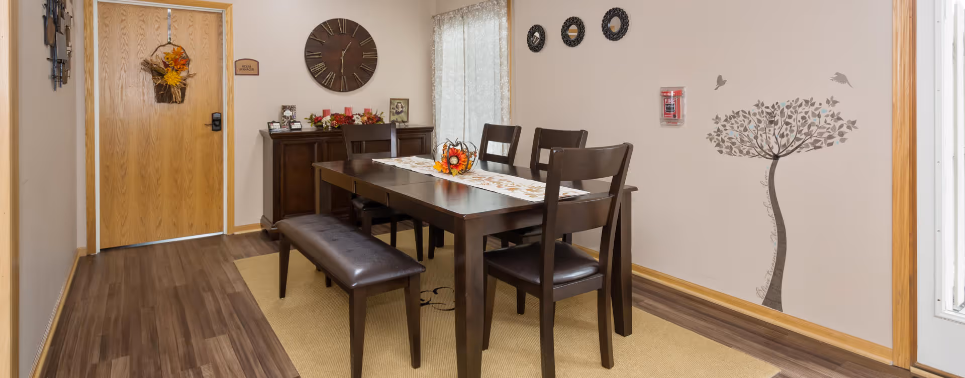 A dining area with a dark wooden table, four chairs, and a bench on one side. The table has a decorative runner and a centerpiece with autumn-themed decor. There is a wooden cabinet against the wall with framed photos and seasonal decorations. A large clock hangs above the cabinet. The walls are light-colored with a tree decal and three circular wall decorations. A door with a fall wreath is visible on the left side, and a window with sheer curtains is on the right.
