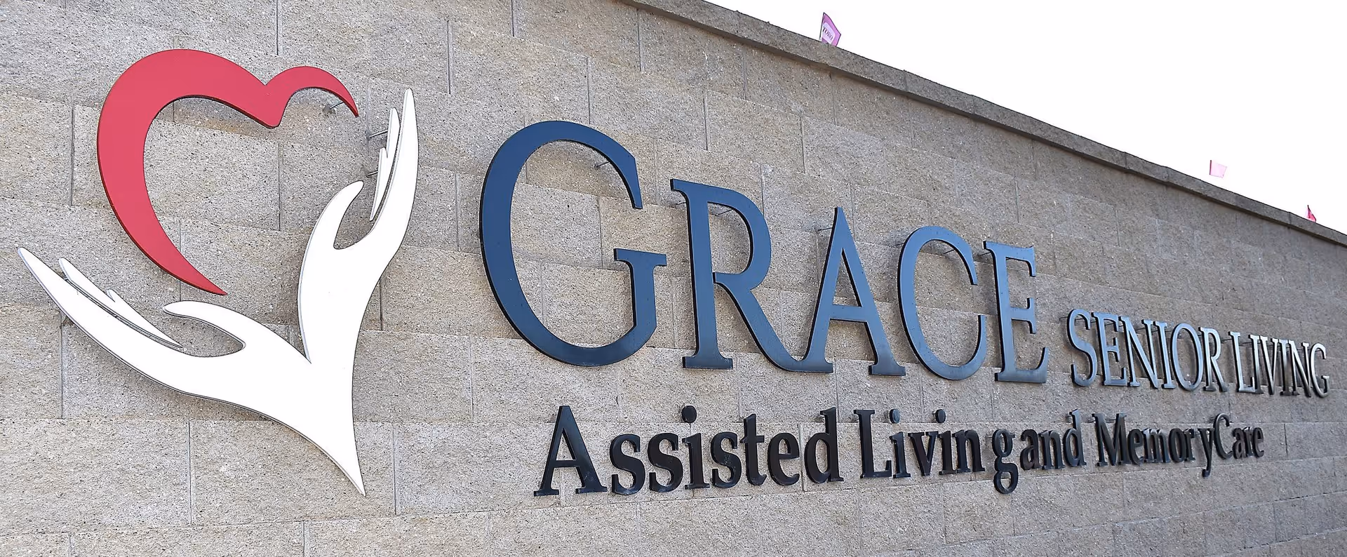 A stone wall with a large sign that reads 'GRACE SENIOR LIVING Assisted Living and Memory Care' alongside a logo featuring two white hands holding a red heart shape.