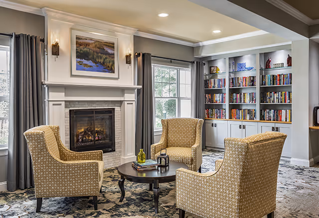 A cozy living room area with three patterned armchairs arranged around a round wooden coffee table. Behind the chairs is a white fireplace with a painting above it and two wall sconces on either side. Large windows with gray curtains allow natural light to fill the room. To the right, there is a built-in bookshelf filled with books and decorative items.