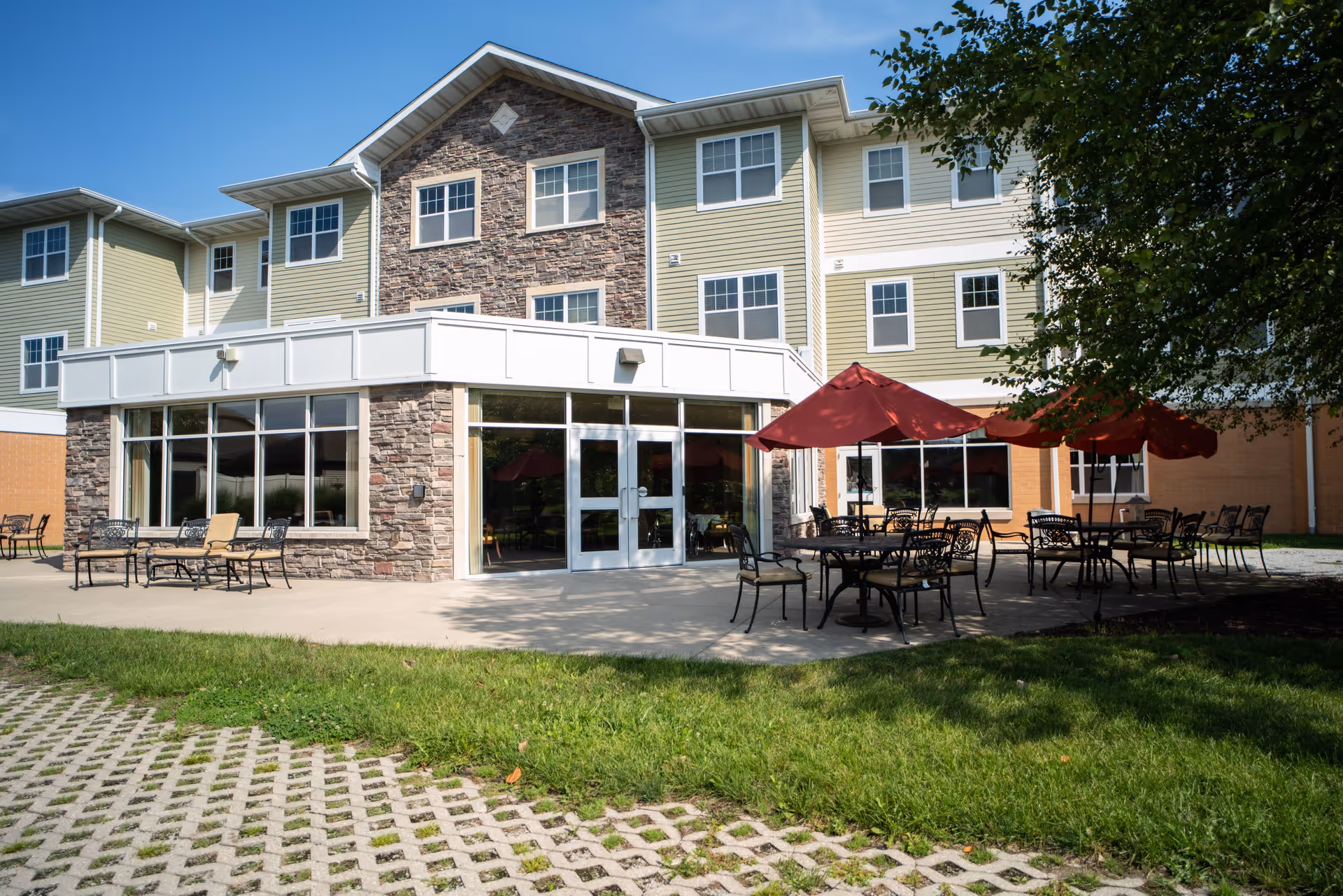 Outdoor patio area of a senior living facility with several black metal tables and chairs, some shaded by red umbrellas, adjacent to a three-story building with stone and beige siding under a clear blue sky.