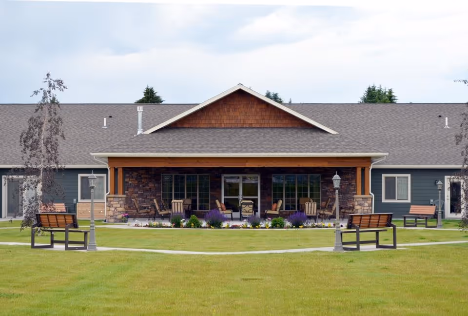 Exterior view of Timbercreek Village Community showing a single-story building with a covered porch area furnished with chairs and tables. The building has a gray roof, stone and wood accents, and is surrounded by a well-maintained lawn with benches and lampposts along a paved walkway.