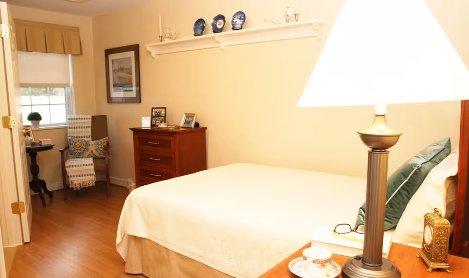 A cozy bedroom with a neatly made bed covered in a light-colored quilt. There is a wooden nightstand with a lamp, a teacup and saucer, a pair of glasses, and a small clock. In the corner, there is a wooden chair with a cushion and a throw blanket, next to a small round table with a potted plant. A wooden dresser with framed photos and decorative items is against the wall, which also features a shelf with decorative plates and candles. The room has warm lighting and a window with a valance and roller shade.