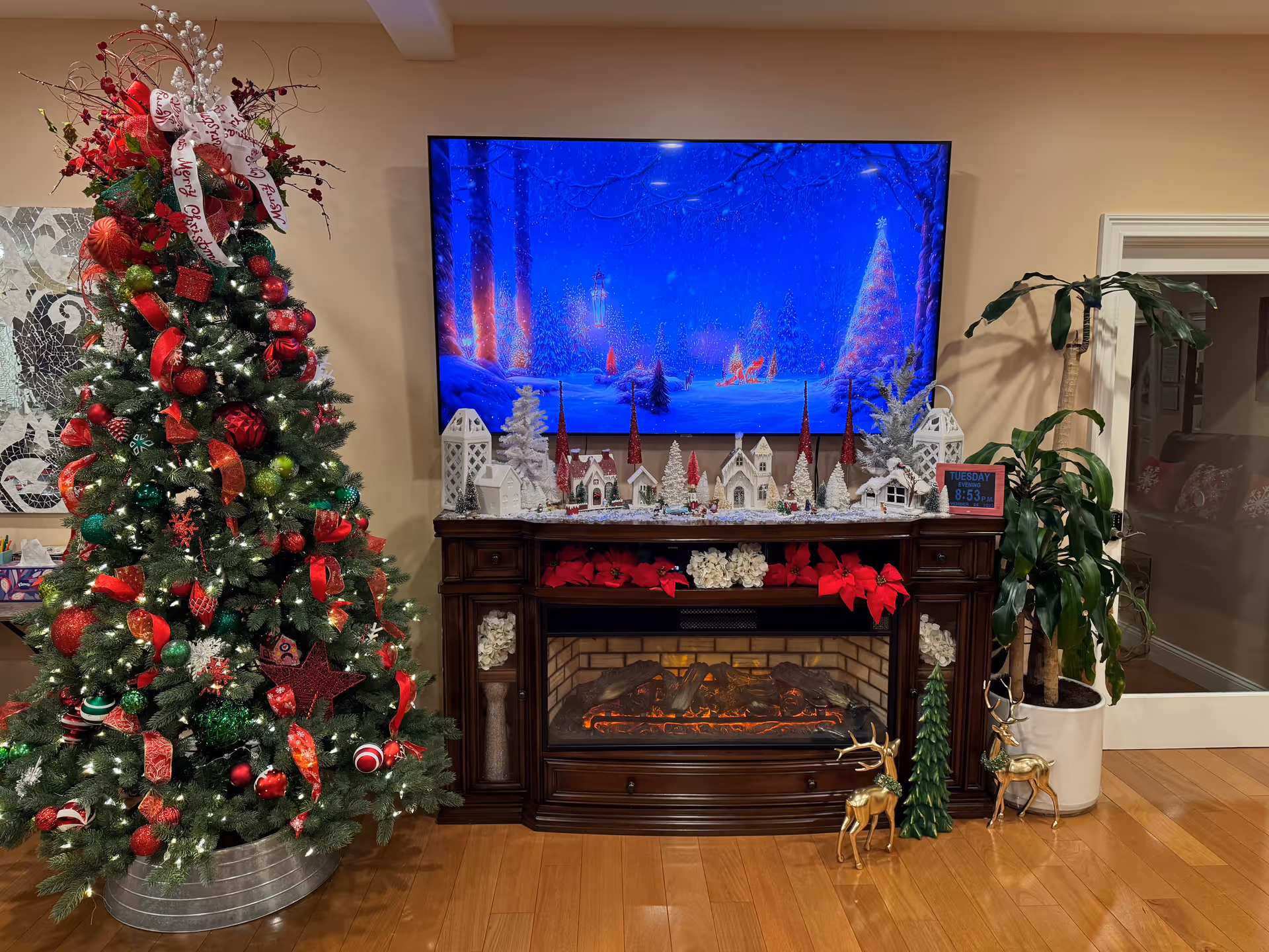 Festively decorated living room corner with a Christmas tree, TV showing a winter scene above a mantel village and an electric fireplace.