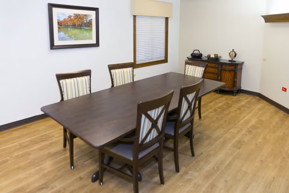 A dining room with a dark wooden rectangular table surrounded by six chairs with striped cushions. There is a window with blinds on the wall behind the table, a framed picture of a lake and trees on the adjacent wall, and a wooden sideboard with decorative items in the corner. The floor is light wood, and the walls are painted white.