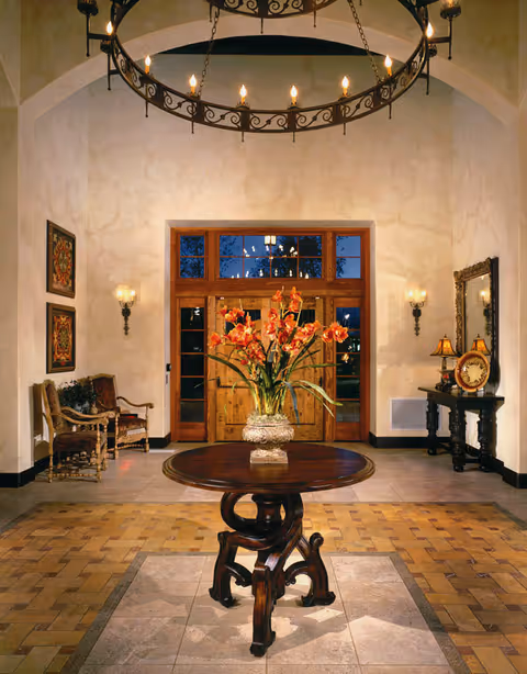 A warmly lit entrance hall with a round wooden table in the center holding a vase of orange flowers. The background features a large wooden door with glass panels, wall sconces with candles on either side, two chairs with ornate wooden frames on the left, and a console table with a mirror and lamps on the right. A large circular chandelier hangs from the ceiling.