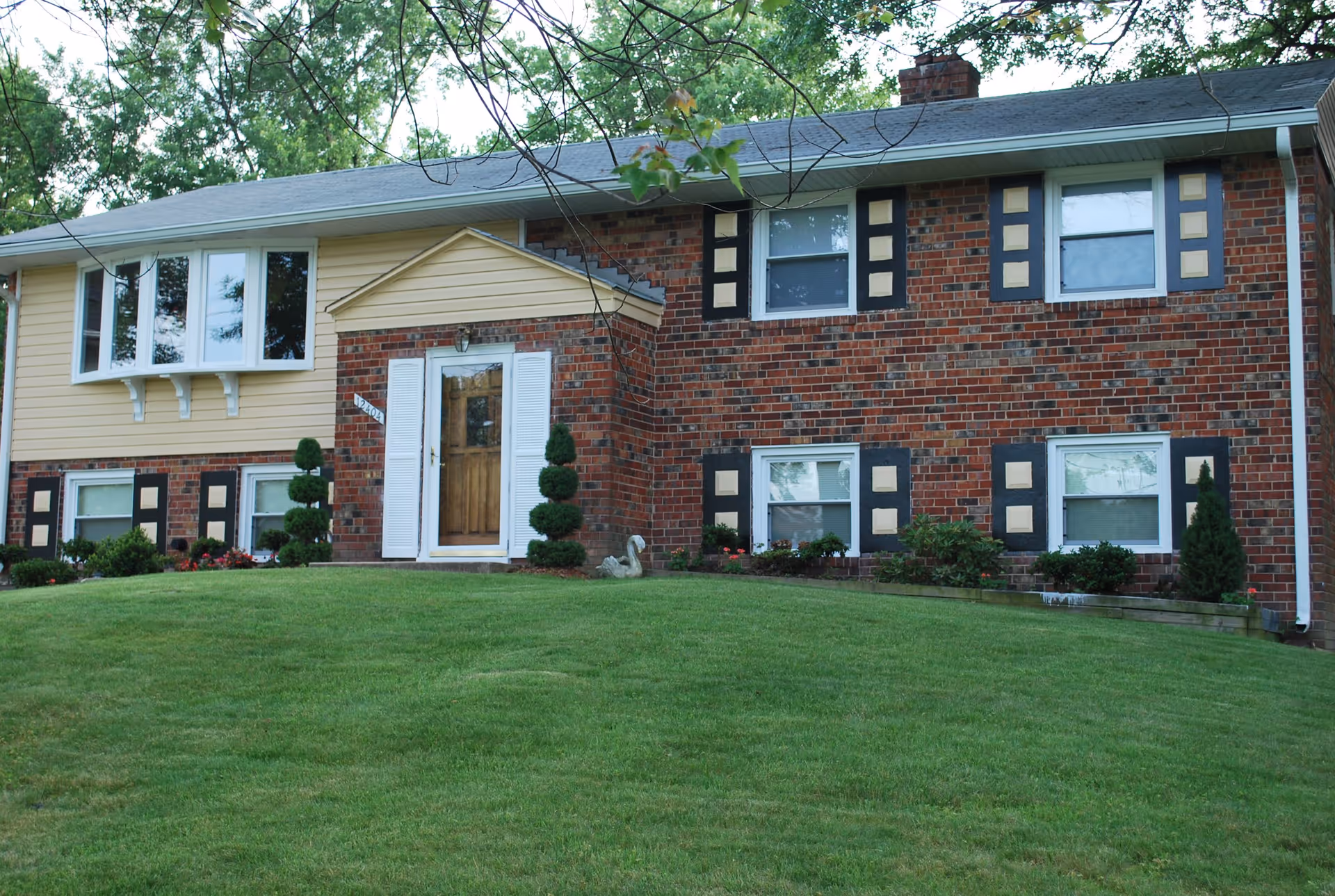 Exterior view of a two-story residential building with a brick and yellow siding facade, white-framed windows, a wooden front door with white shutters, and a well-maintained green lawn with shrubs and small trees.