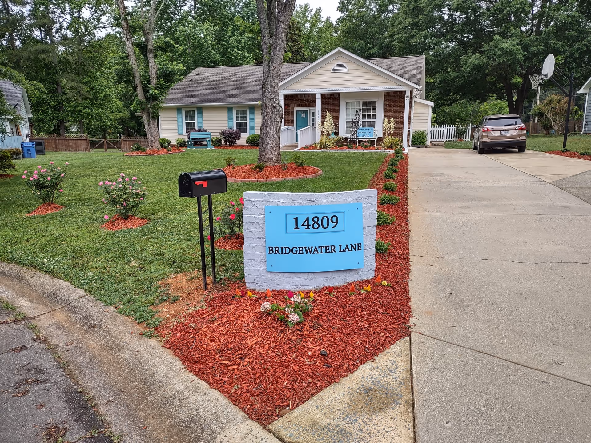 A single-story house with a brick and beige exterior, a front porch with white columns, and a driveway with a parked car. The lawn is well-maintained with green grass, flower beds with red mulch, and small bushes. A black mailbox stands near the curb next to a white brick sign with a blue plaque displaying the address '14809 Bridgewater Lane'. Trees and neighboring houses are visible in the background.