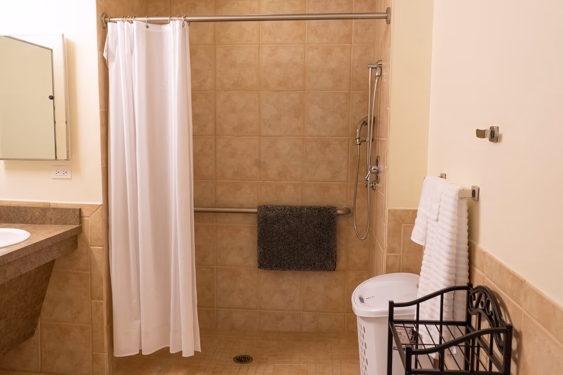 A bathroom with beige tiled walls and floor, featuring a walk-in shower with a white shower curtain, a handheld showerhead, and a grab bar with a dark towel hanging on it. To the right, there is a white laundry basket, a black metal rack, and two white towels hanging on a towel rack. On the left side, a countertop with a sink and a mirror above it is partially visible.