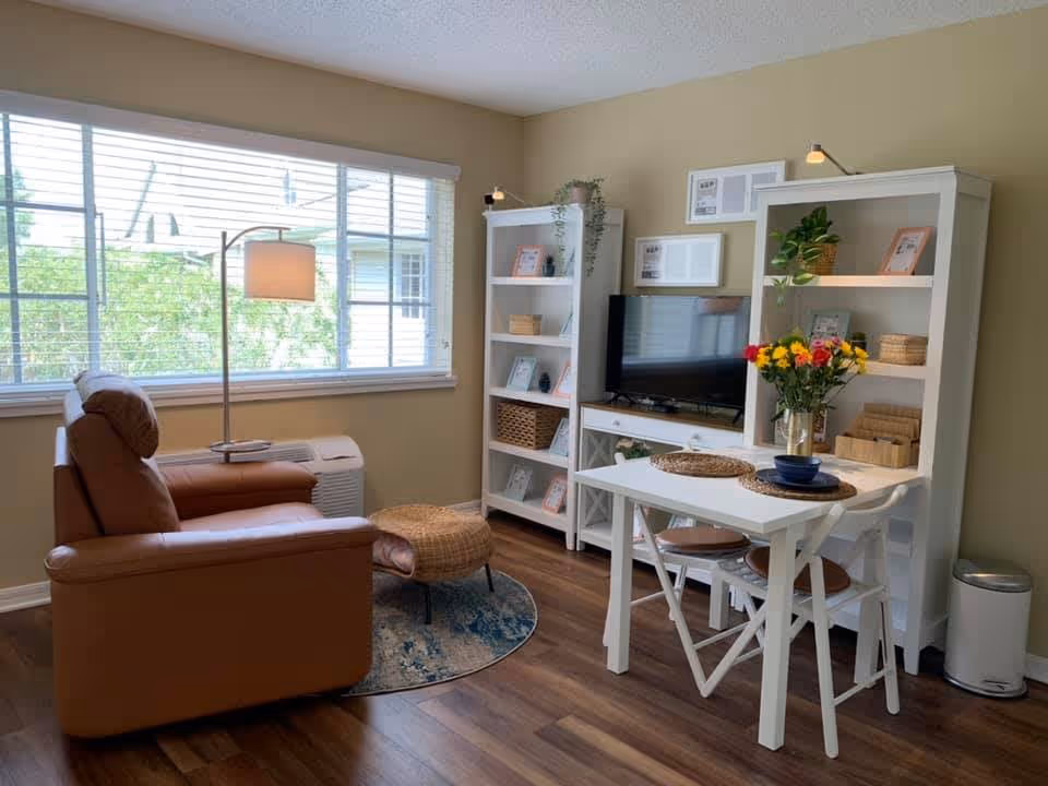 A cozy living room with a large window letting in natural light. The room features a brown leather armchair with a matching ottoman on a round rug, a white floor lamp, two white bookshelves filled with framed photos, plants, and baskets, a flat-screen TV on a white TV stand, and a small white dining table with two chairs. A vase with colorful flowers is on the table, and the floor is wooden.
