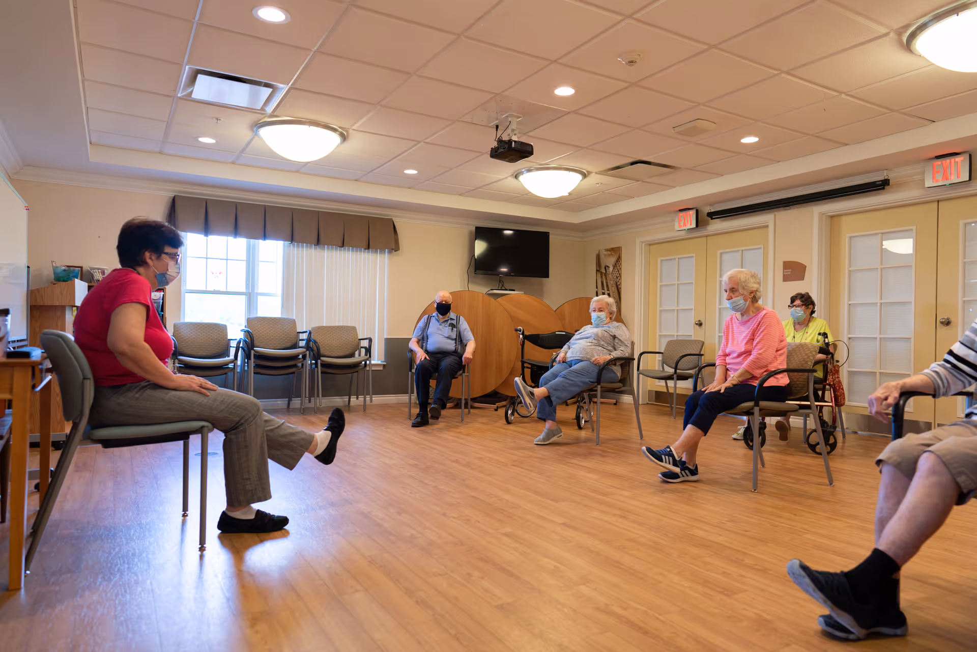 A group of elderly people sitting in chairs spaced apart in a large room with wooden flooring, participating in a seated exercise or activity led by an instructor wearing a red shirt and gray pants. Everyone is wearing face masks. The room has beige walls, a window with blinds, stacked chairs, a wall-mounted TV, and exit signs above doors.