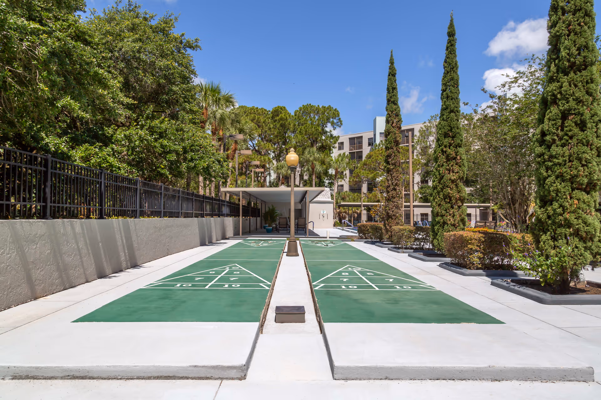Outdoor shuffleboard courts with green playing surfaces and white markings, surrounded by trees, bushes, and a building in the background under a blue sky with some clouds.