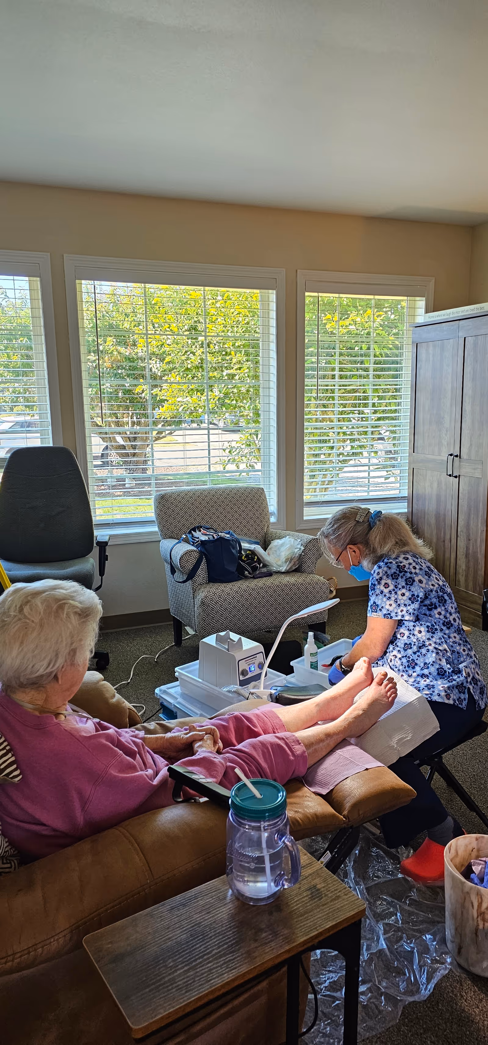 An elderly woman is seated on a brown recliner chair receiving foot care from a caregiver wearing a blue floral shirt and a face mask in a well-lit room with large windows. The room has a gray armchair, a wooden cabinet, and a small table with a water bottle on it.
