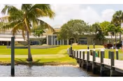 View of a waterfront area with a wooden dock extending over the water, palm trees, green grass, and a large building in the background under a partly cloudy sky.