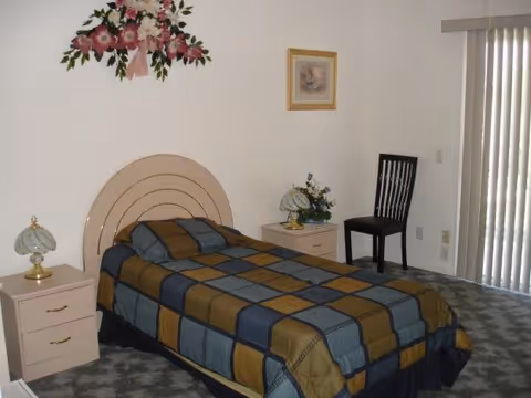 A bedroom with a single bed covered in a blue, brown, and green checkered bedspread. There are two beige nightstands on either side of the bed, each with a decorative lamp. A black chair is placed near vertical blinds covering a window or sliding door. The walls are white with a floral wall decoration above the bed and a framed picture on the wall.