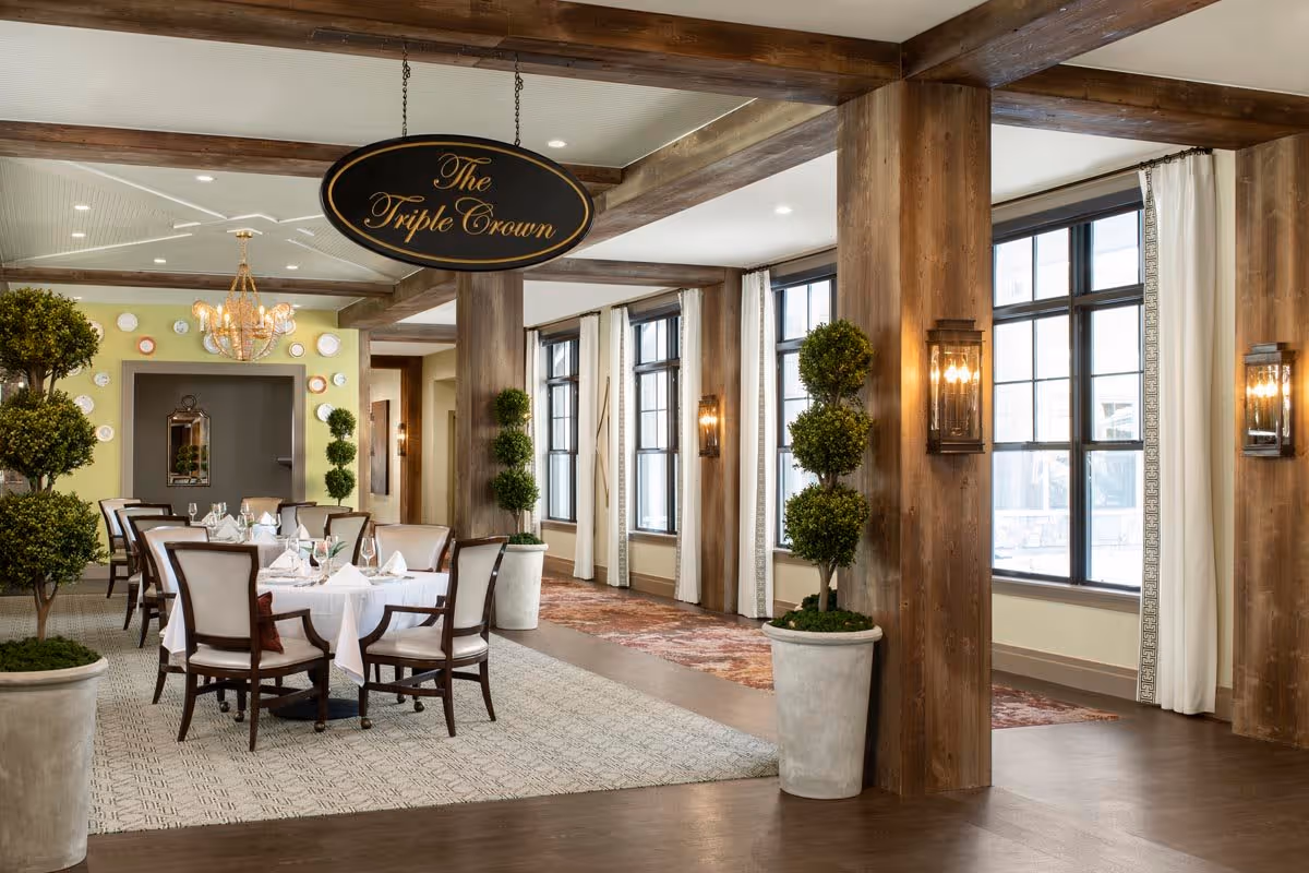 A dining room with a table set for six people with white tablecloths, napkins, and glassware. The room features large windows with white curtains, wooden beams and columns, potted topiary plants, and a chandelier. A sign hanging from the ceiling reads 'The Triple Crown'.