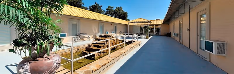 Outdoor courtyard area at Vincent Victoria Village Assisted Living featuring a stone water fountain with a metal railing, potted plants, and beige building walls with doors and windows under a clear blue sky.
