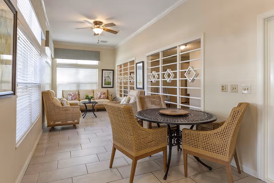 A bright and airy living room area with large windows covered by blinds, a ceiling fan with light, and light-colored tiled flooring. The room features a seating area with a beige sofa, two wicker armchairs with cushions, a small black coffee table with a plant, and a round black metal table with four wicker chairs. The walls are light-colored with decorative white lattice panels and framed artwork.