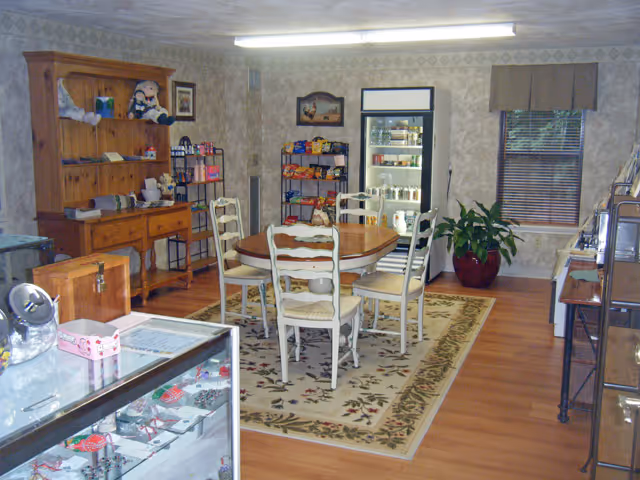 A cozy dining area with a wooden round table and four white chairs on a floral rug. The room has wooden flooring and wallpapered walls. There is a wooden hutch with various items on it, a refrigerated display case with snacks and drinks, a potted plant near a window with blinds, and shelves stocked with snacks and other items.