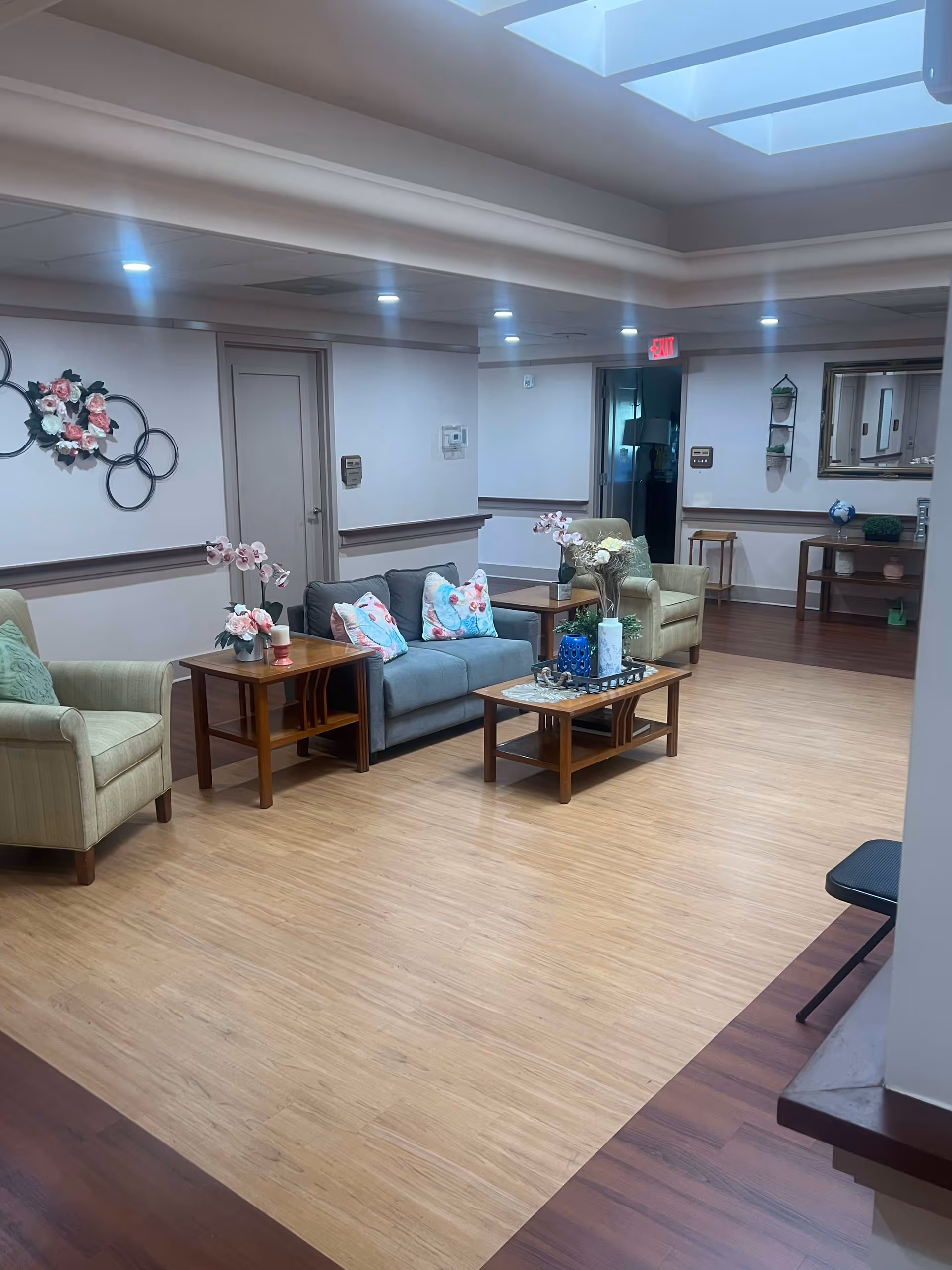 A cozy senior living community lounge area with a gray sofa adorned with floral pillows, two beige armchairs, wooden coffee and side tables decorated with flower vases, and a wall decoration featuring circular metal rings with pink flowers. The room has a light wood floor, recessed ceiling lights, and a skylight.